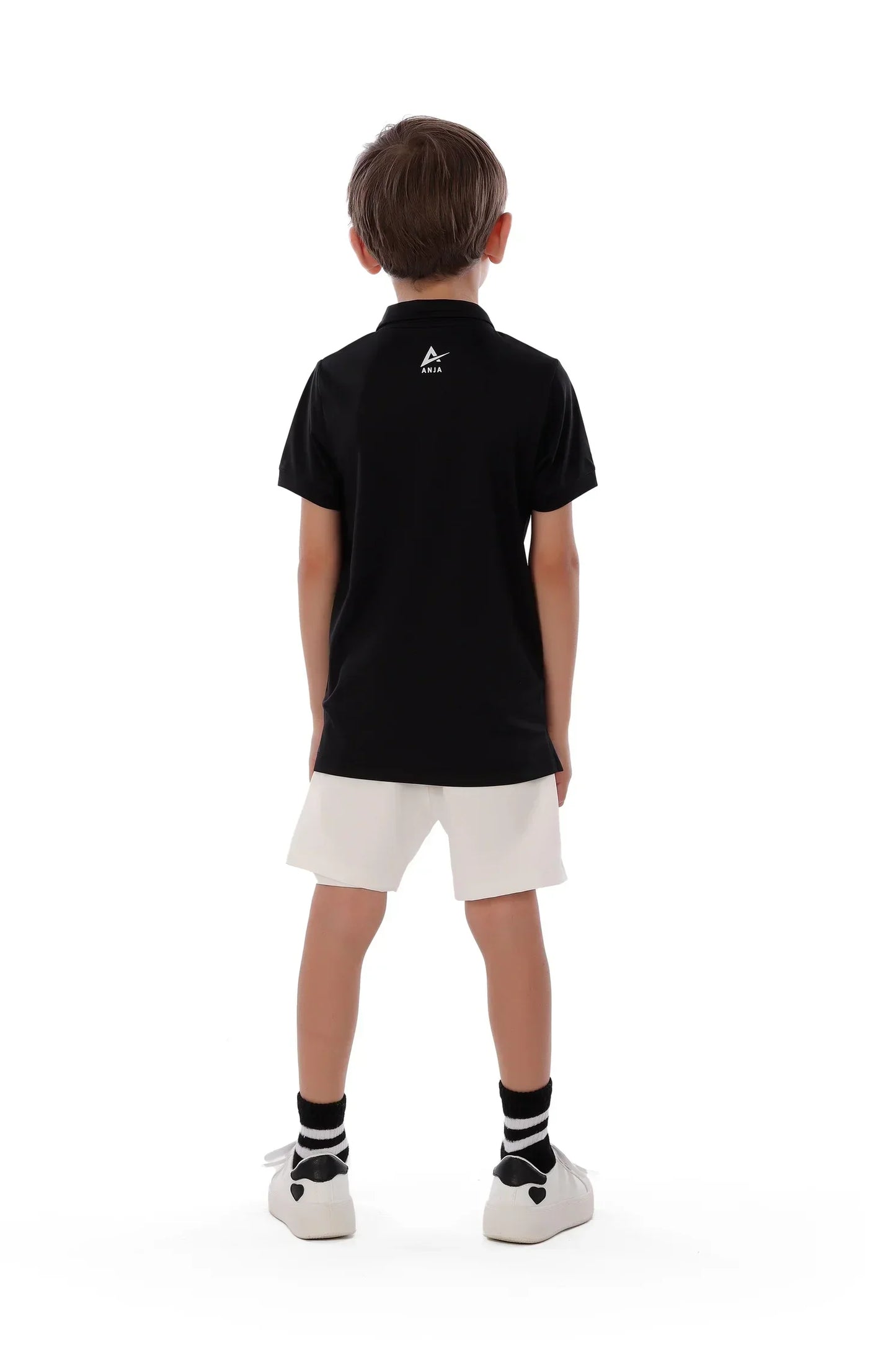 Child wearing a black polo shirt with a logo on the back, white shorts, and black socks with white stripes.