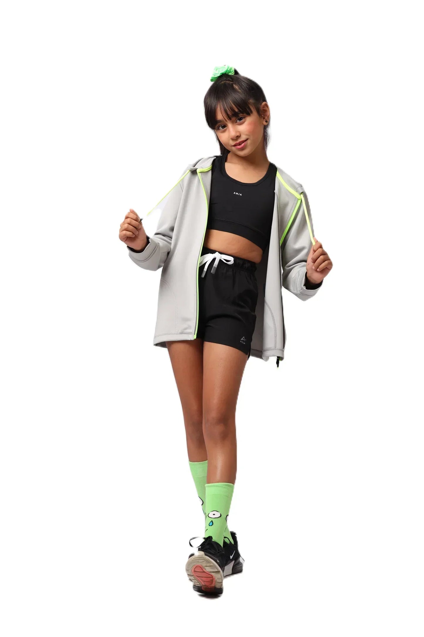 girl wearing a gray jacket, black athletic top, and shorts with green socks on a white background