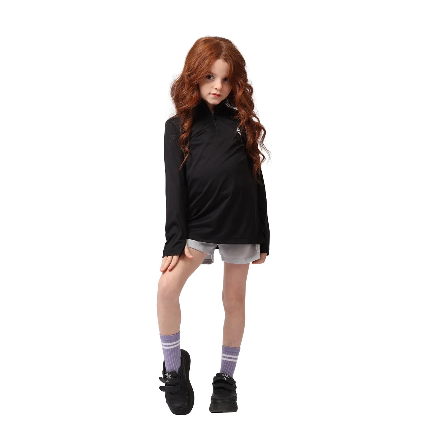 Young girl wearing a black long-sleeve shirt, gray shorts, and black shoes on a white background