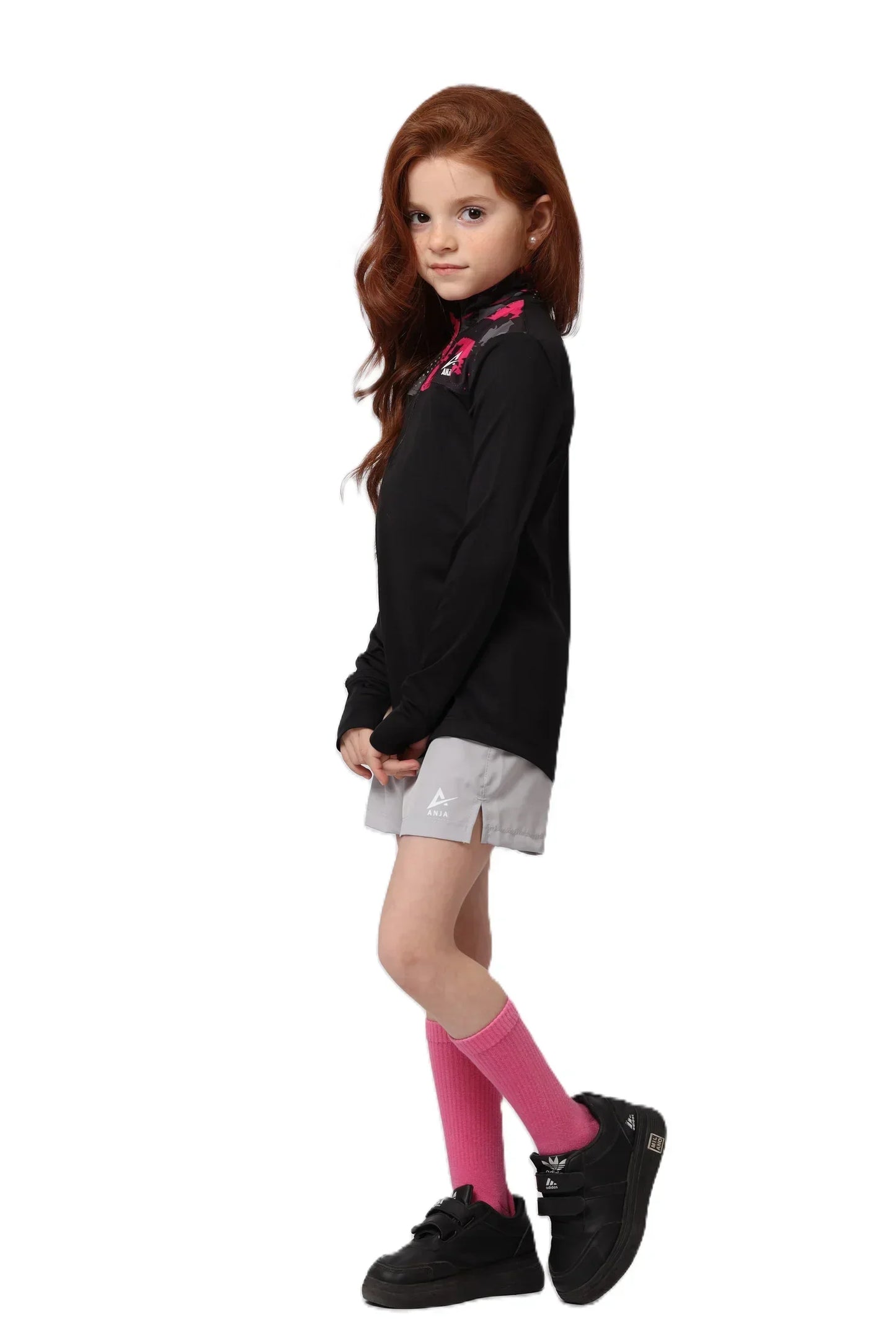 Young girl wearing a black shirt with pink floral details, white shorts, pink socks, and black shoes on a white background