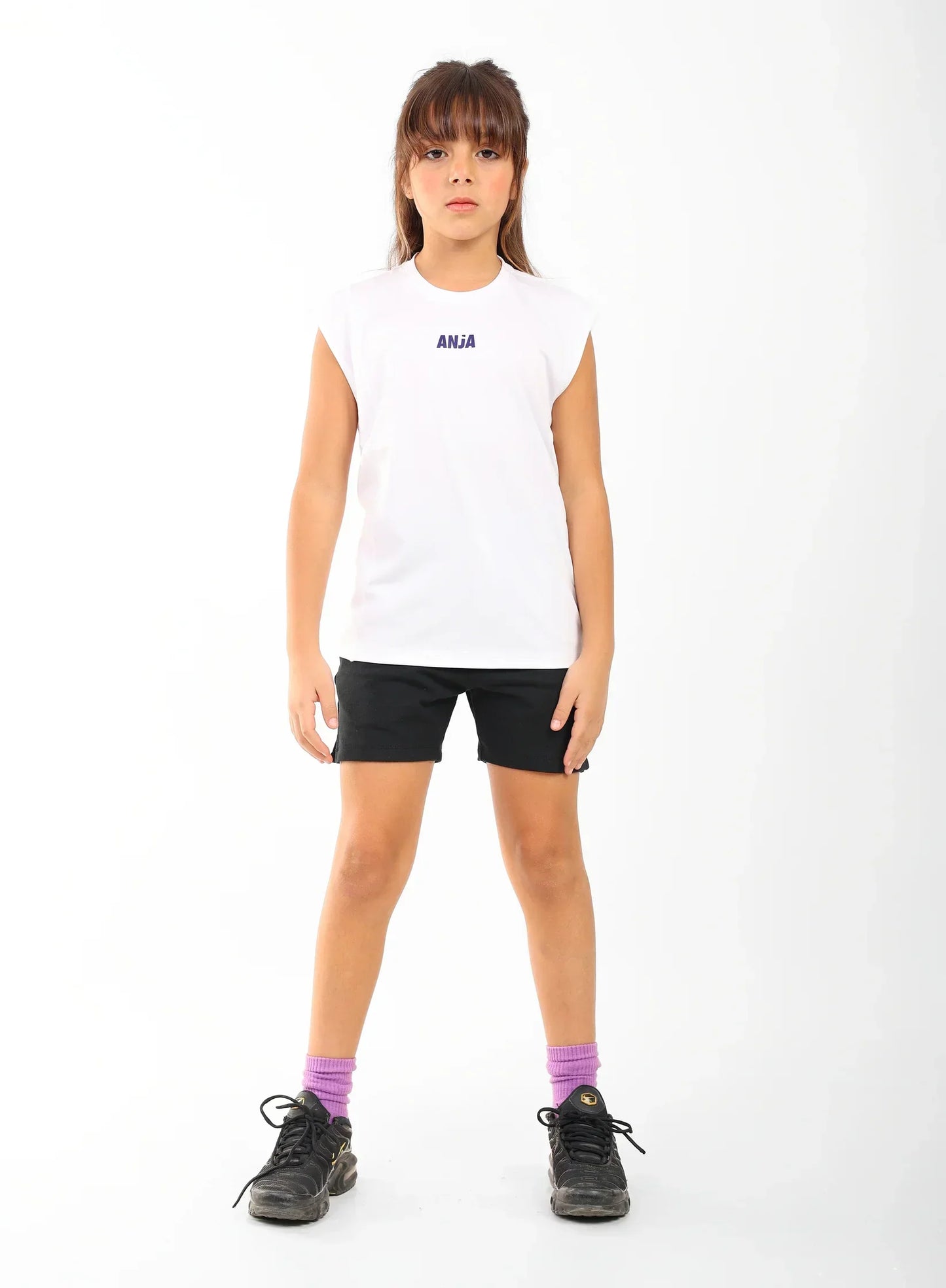 Young girl wearing a white sleeveless shirt with a logo, black shorts, and purple socks on a white background